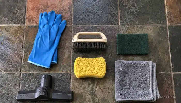 Slate floor cleaning tools including grout brush, scrubbing pad, gloves and wet vacuum nozzle arranged on a riven slate surface